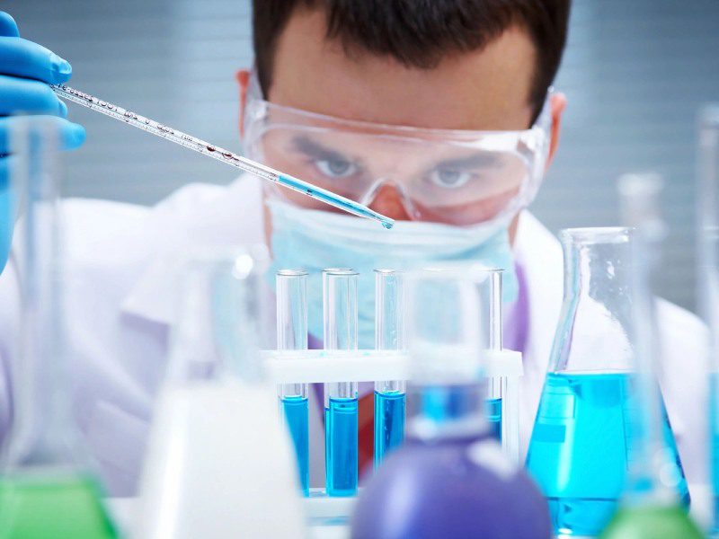 A man in lab coat and goggles looking at blue liquid.